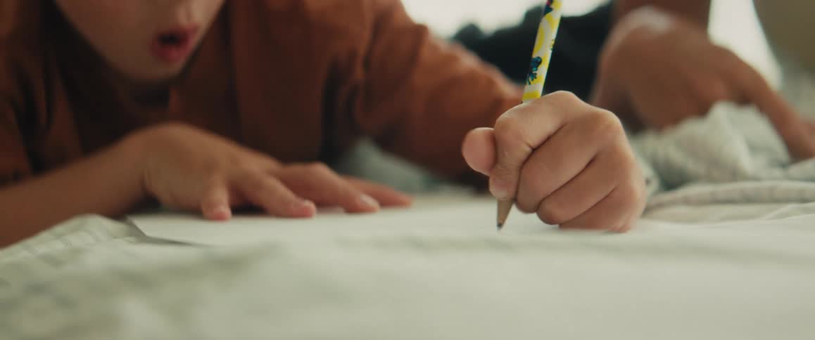 A focused young boy lies on his stomach, concentrating intently as he draws with a pencil on a piece of paper