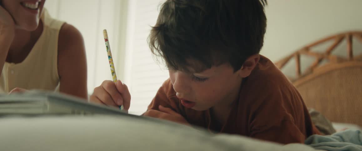 A loving mother smiles as she helps her young son with his homework while they relax on a bed at home