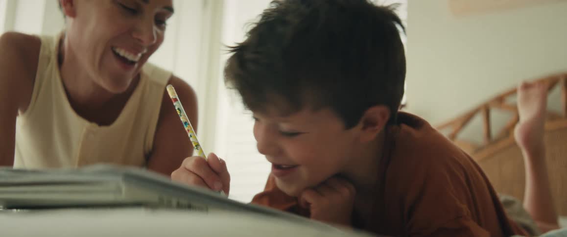 A loving mother smiles as she helps her young son with his homework while they relax on a bed at home