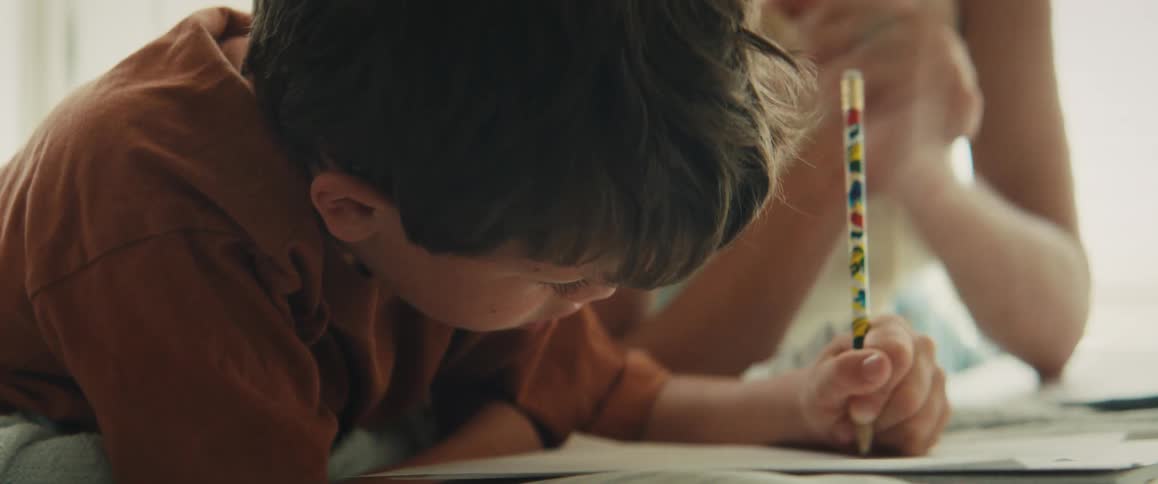 A loving mother gently encourages her young son as he lies down and carefully draws with a pencil at home
