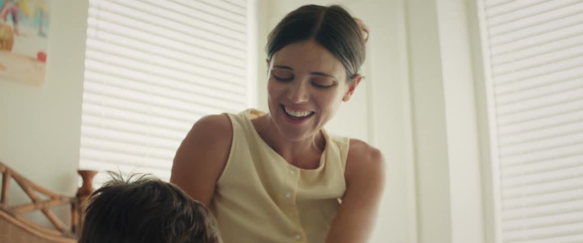 A mother affectionately watches her son as he happily draws with a pencil on the bed
