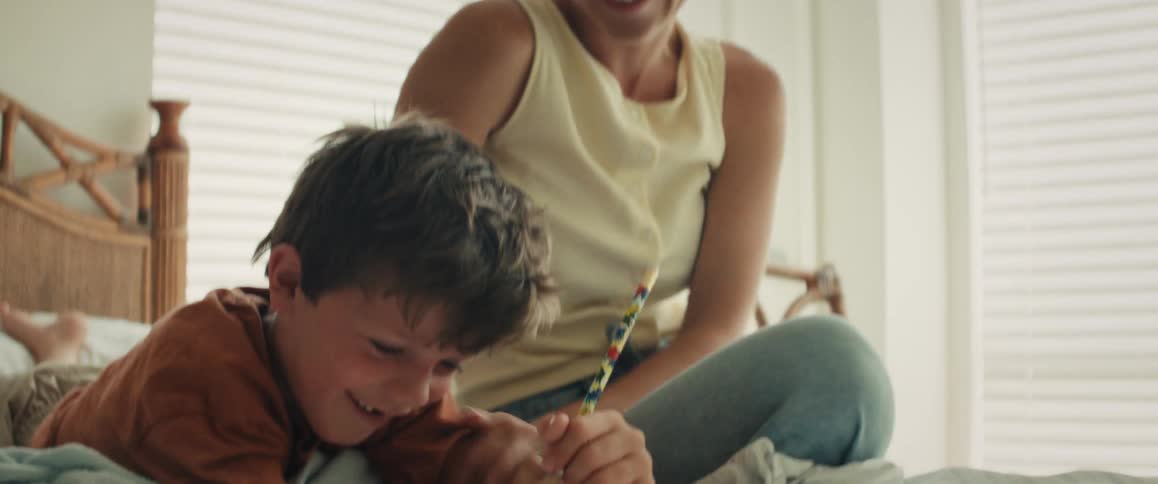 A mother affectionately watches her son as he happily draws with a pencil on the bed