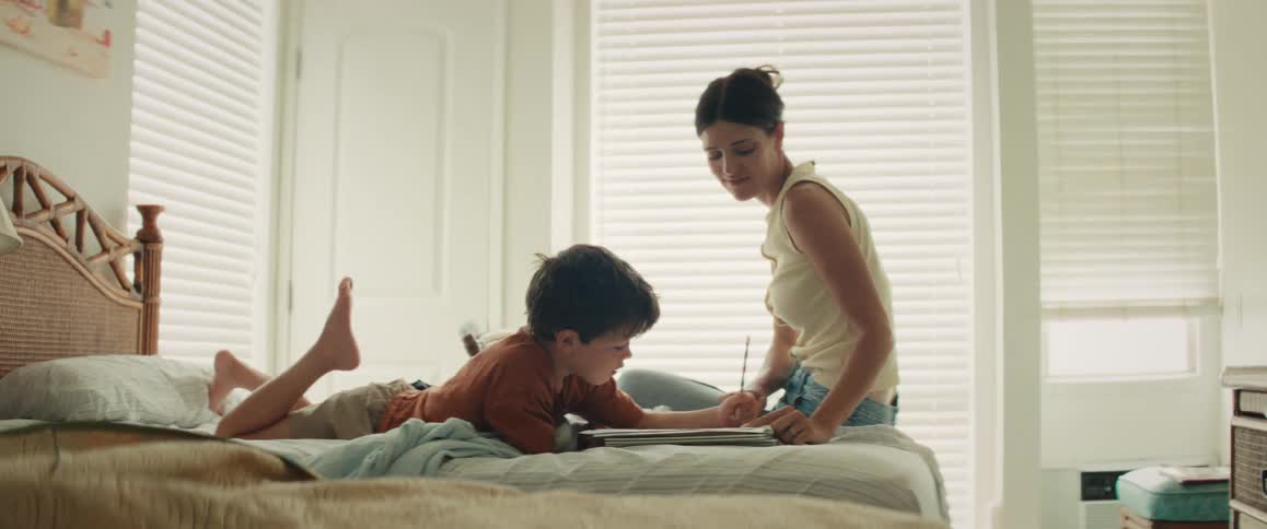 A smiling mother helps her young son with his drawing as they spend quality time together on a bed in a bright bedroom