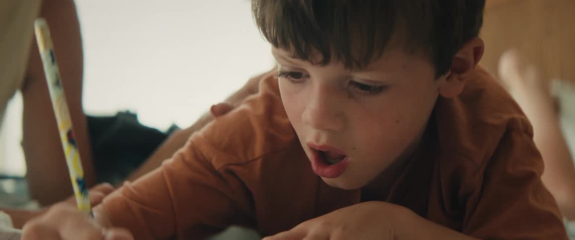A close-up of a young boy intensely focused on drawing with a pencil while his mother's gentle hand rests on his shoulder
