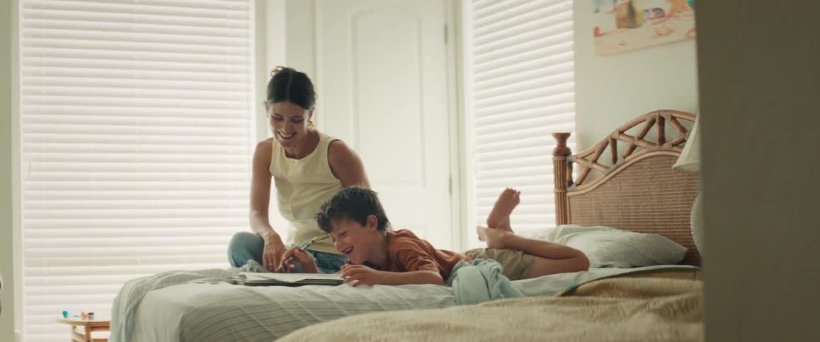 A mother and son laugh together while reading a book in a cozy, sunlit bedroom