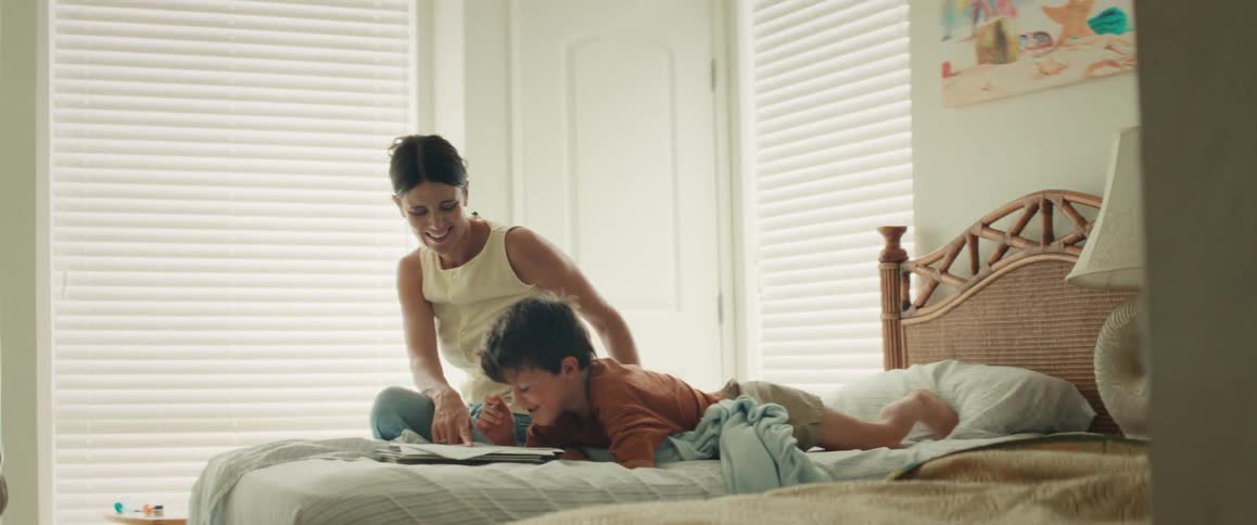 A mother and son laugh together while reading a book in a cozy, sunlit bedroom