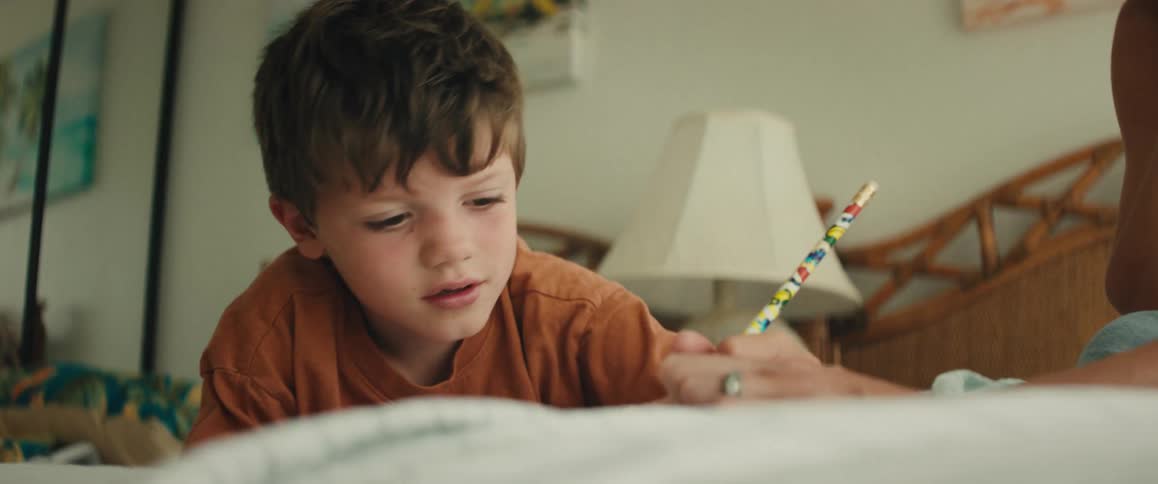 A concentrated young boy lies on his bed and carefully draws with a pencil as his mother helps him