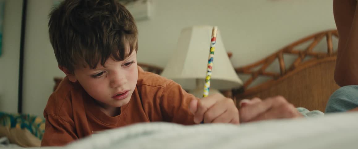 A concentrated young boy lies on his bed and carefully draws with a pencil as his mother helps him