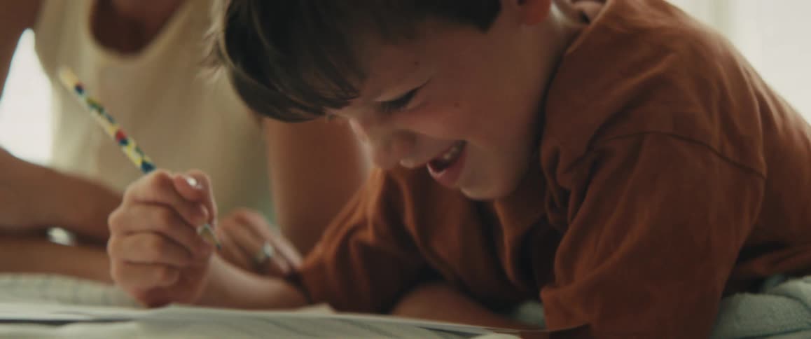 A young boy laughs joyfully while drawing as his mother playfully tickles and kisses him on the forehead