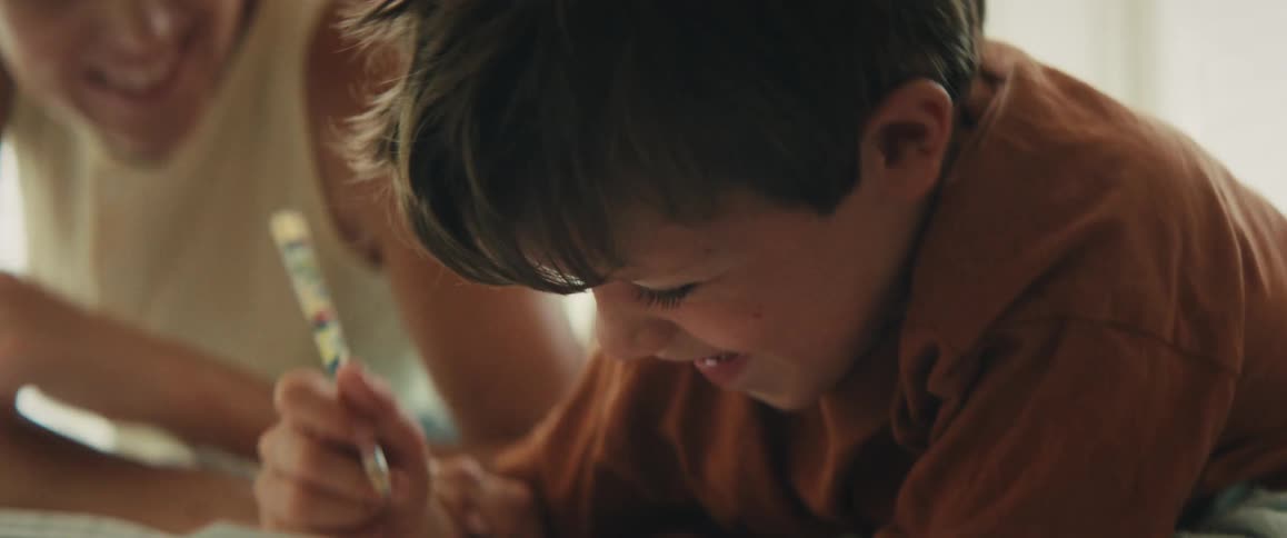 A young boy laughs joyfully while drawing as his mother playfully tickles and kisses him on the forehead