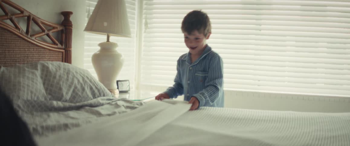 A smiling young boy in blue striped pajamas carefully makes a large bed in a sunlit room in slow motion