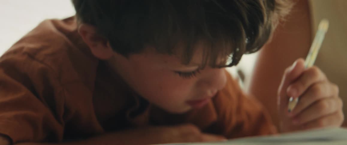 A close-up of a focused young boy wearing a rust-colored shirt as he carefully draws with a pencil