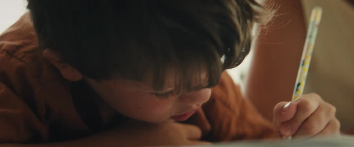 A close-up of a focused young boy wearing a rust-colored shirt as he carefully draws with a pencil