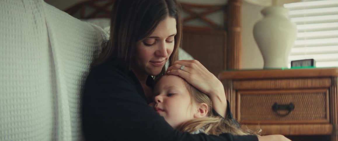 A loving mother tenderly holds her young daughter, gently stroking her hair as they share a quiet moment in a cozy bedroom