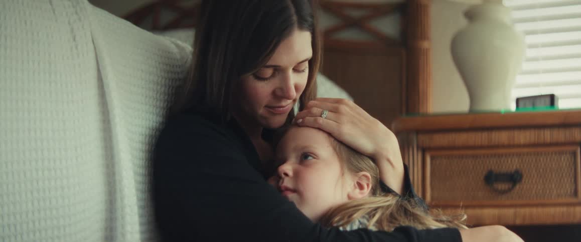 A loving mother tenderly holds her young daughter, gently stroking her hair as they share a quiet moment in a cozy bedroom