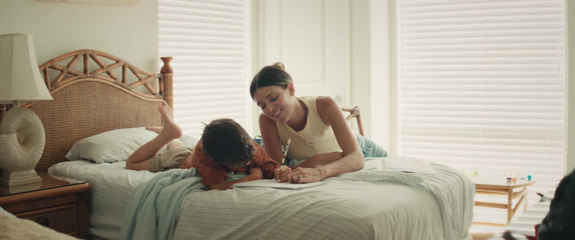 A mother and son spend quality time together, drawing and laughing while relaxing on a bed in a sunlit room