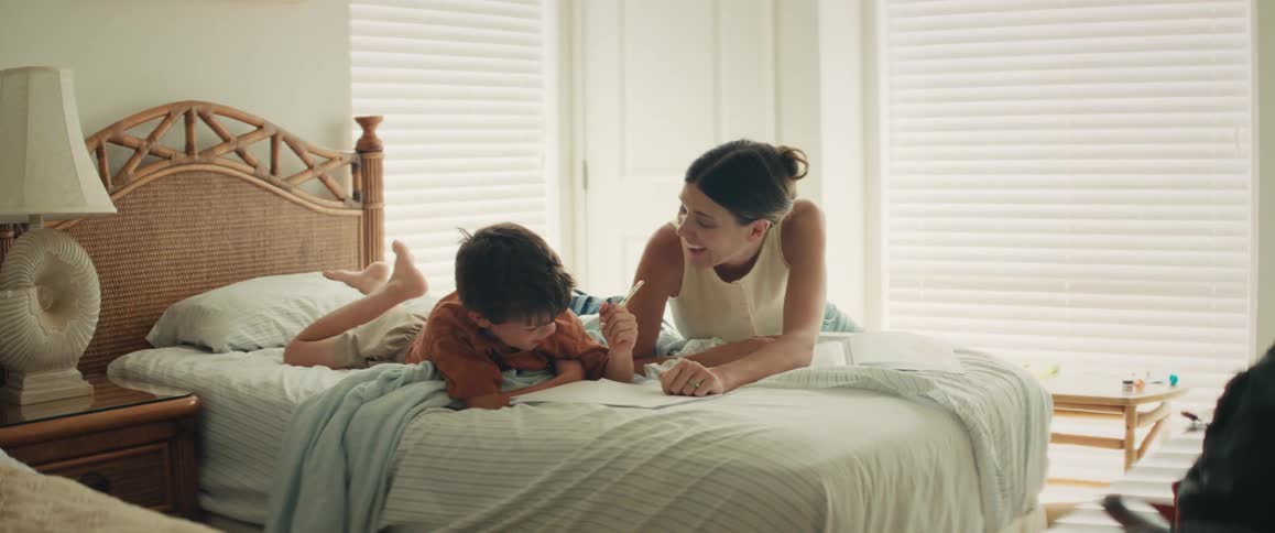 A mother and son spend quality time together, drawing and laughing while relaxing on a bed in a sunlit room