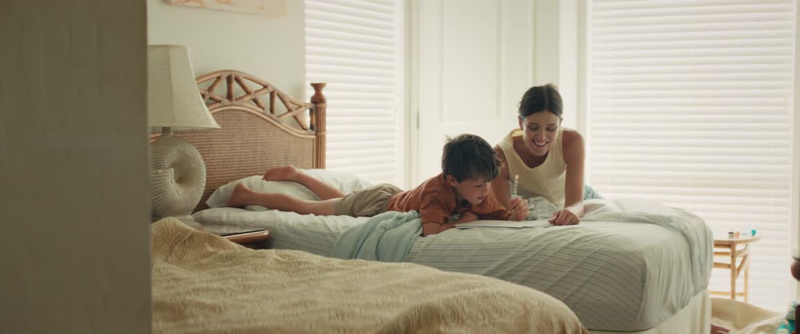 A mother and her young son happily spend time together drawing and laughing while lying on a bed in a bright bedroom