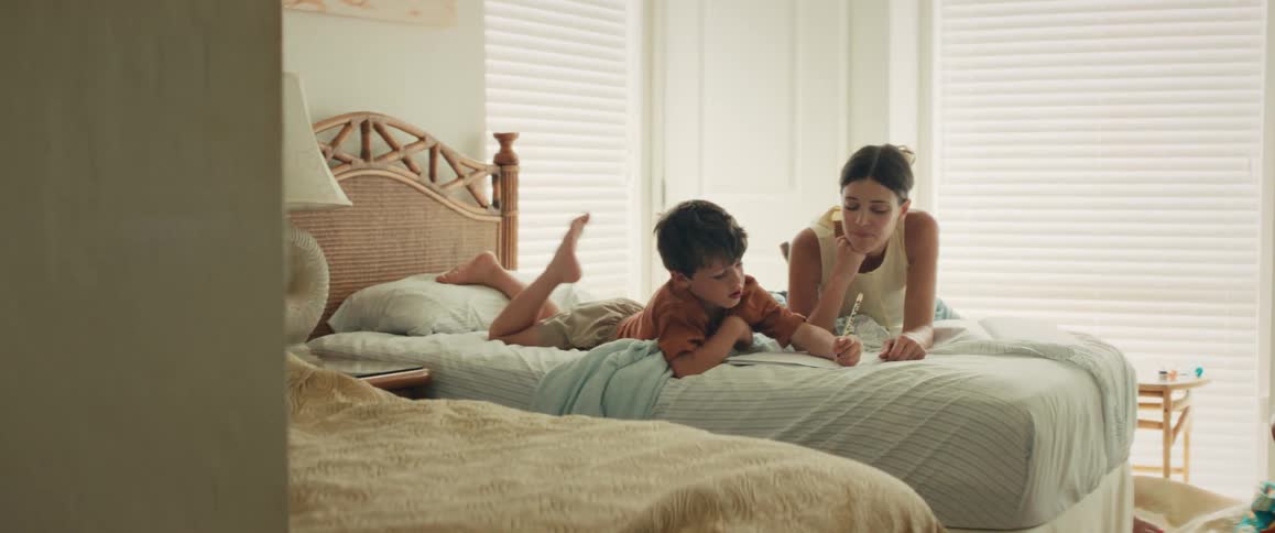 A mother and her young son happily spend time together drawing and laughing while lying on a bed in a bright bedroom