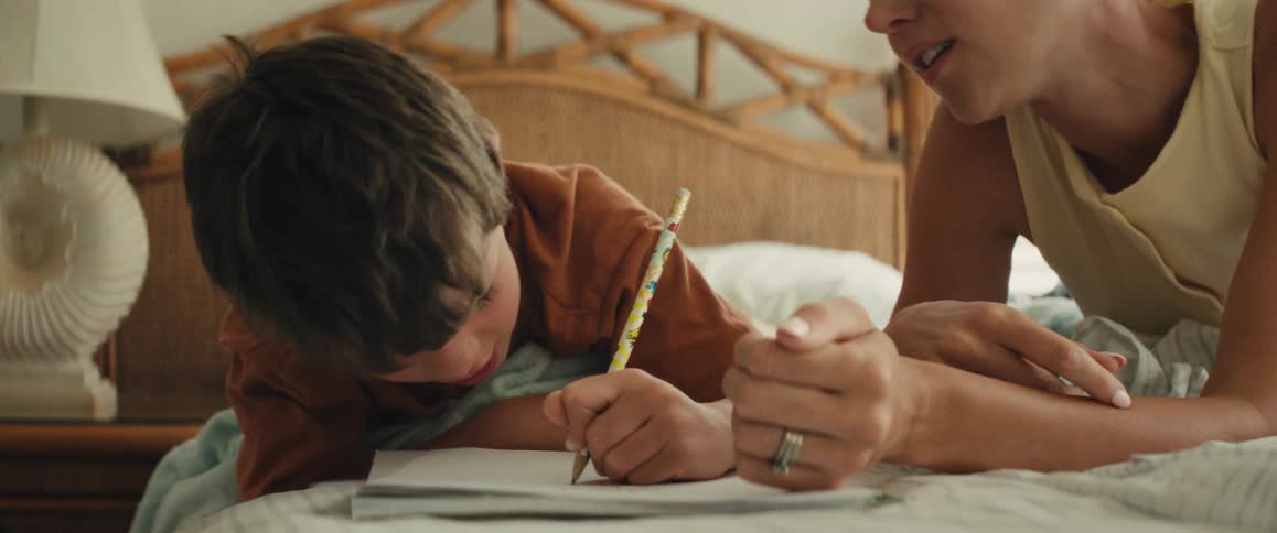 A loving mother guides her young son's hand with a pencil as he learns to write while they share a sweet moment lying on a bed