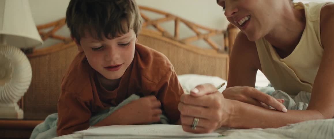 A loving mother guides her young son's hand with a pencil as he learns to write while they share a sweet moment lying on a bed