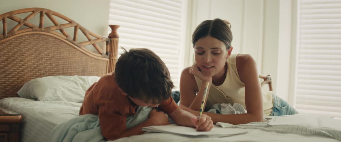 A loving mother patiently helps her young son with drawing in a notebook while they relax together on a bed in a sunlit bedroom