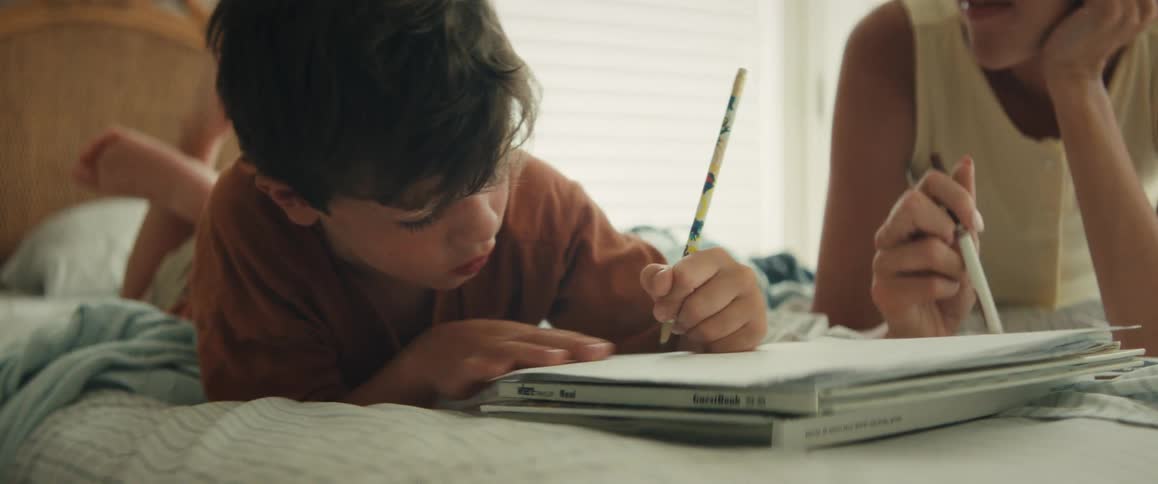 A young boy concentrates while drawing with a pencil in a sketchbook as he lays on a bed next to his mother