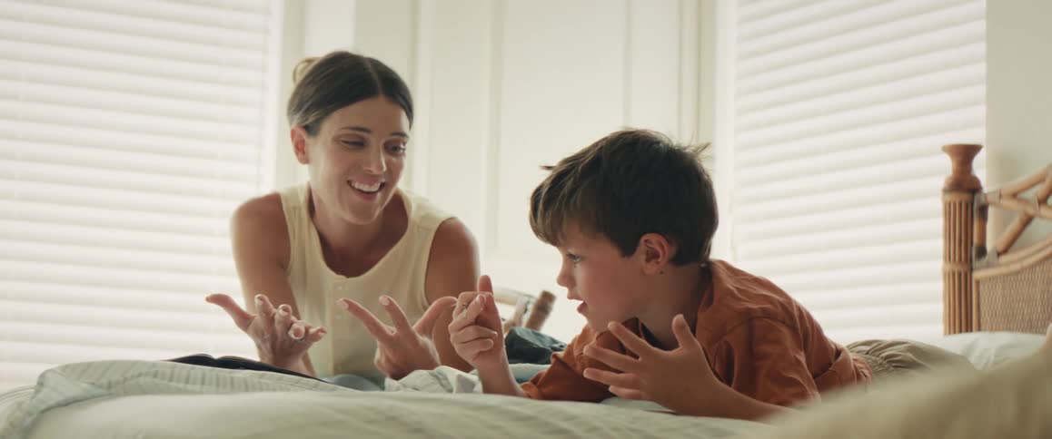 A caring mother helps her young son with his homework, encouraging him as he counts on his fingers while they lie together on the bed in a bright, sunlit room