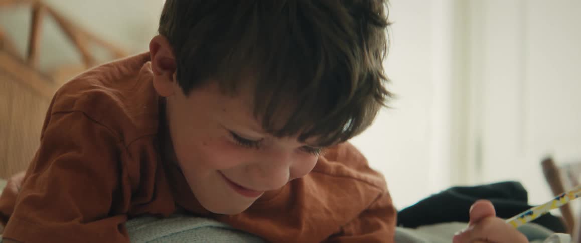 Joyful young boy laughs playfully before concentrating on drawing while lying on his bed