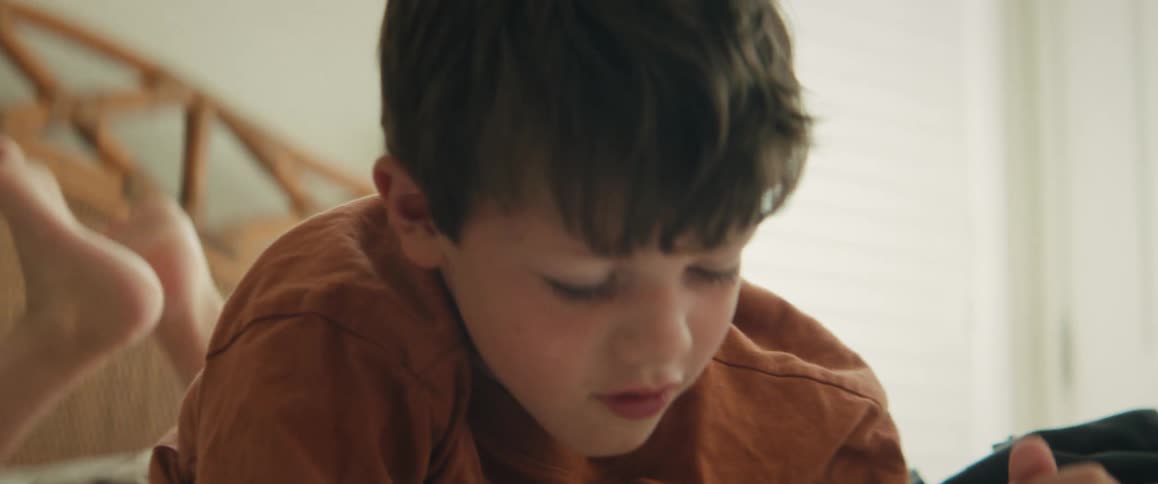 Joyful young boy laughs playfully before concentrating on drawing while lying on his bed