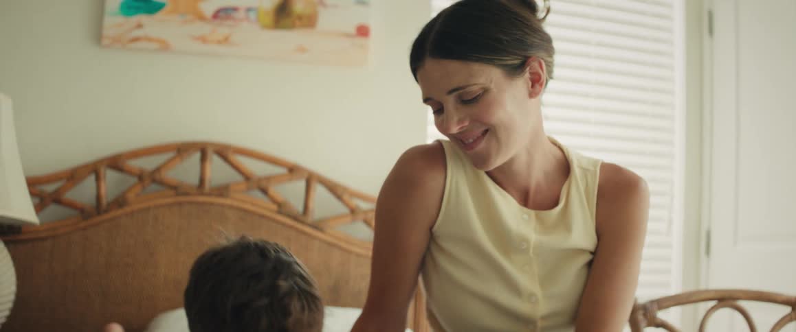 A loving mother smiles as she sits on a bed and watches her young son concentrate on his homework