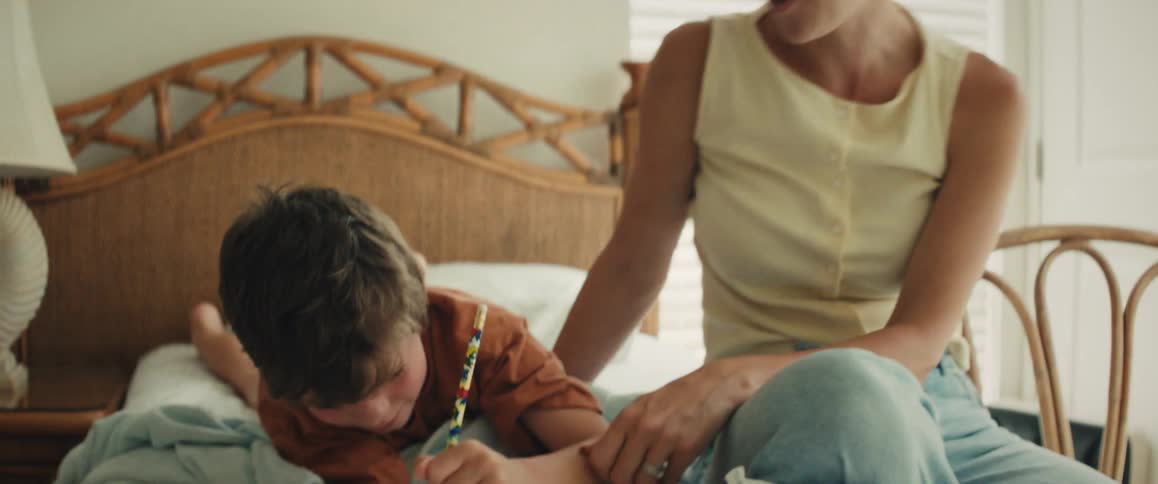 A loving mother smiles as she sits on a bed and watches her young son concentrate on his homework