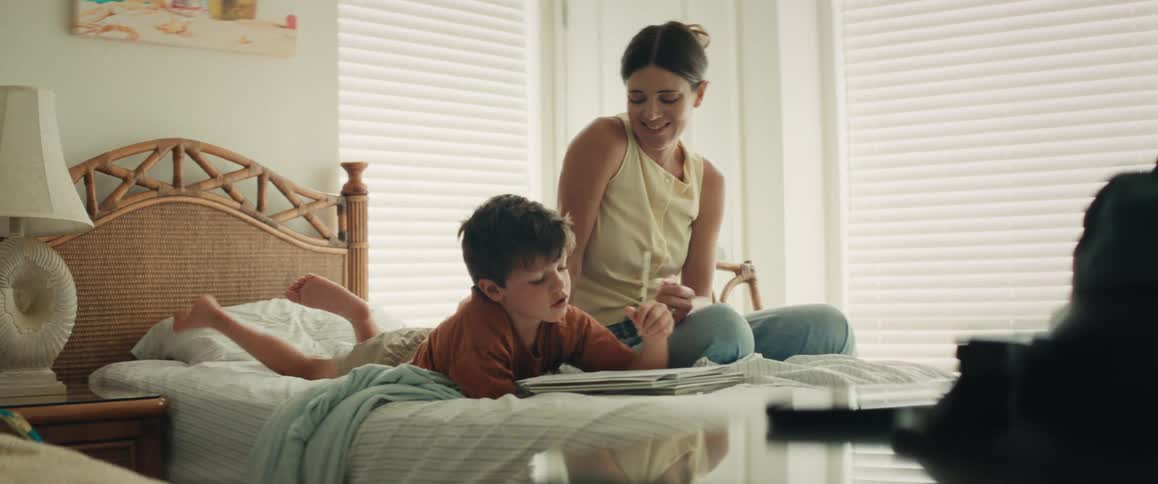 A young boy lies on his stomach on the bed, drawing in a notebook, while his mother sits beside him offering loving encouragement