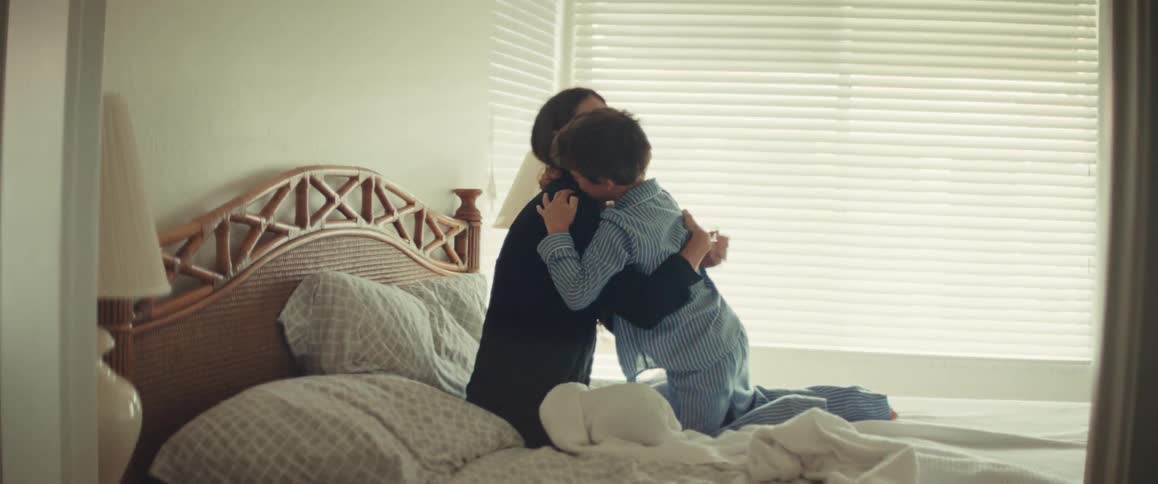 A loving son in pajamas crawls across the bed to give his mother a warm morning hug and kiss in slow motion