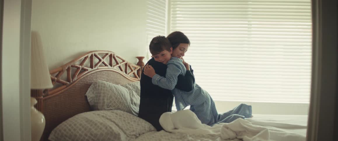 A loving son in pajamas crawls across the bed to give his mother a warm morning hug and kiss in slow motion