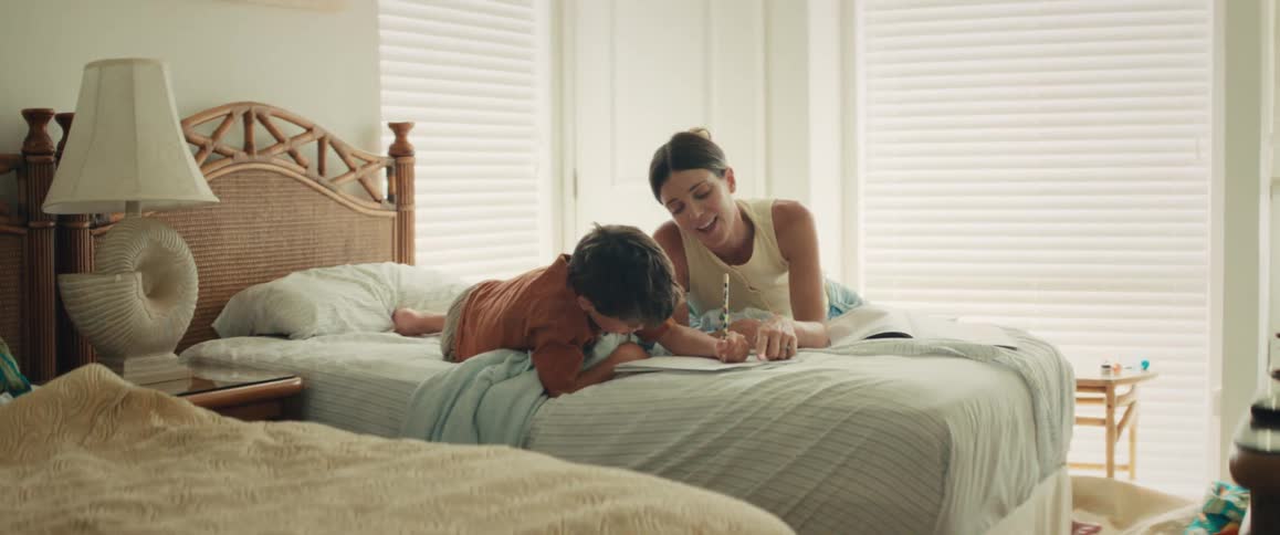 A happy mother teaches her young son how to write in a notebook as they relax together on a bed in a sunlit bedroom