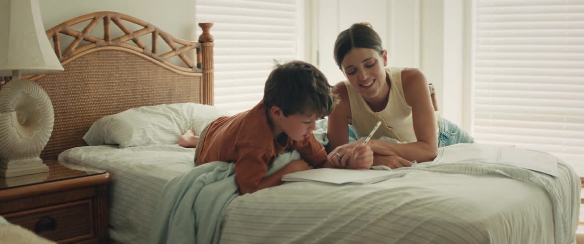 A happy mother teaches her young son how to write in a notebook as they relax together on a bed in a sunlit bedroom