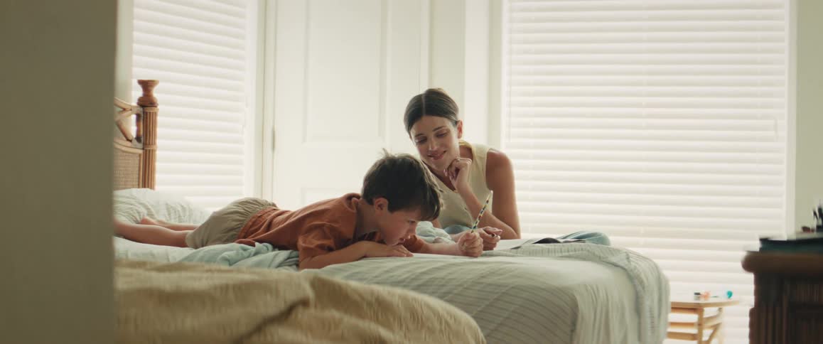 A loving mother helps her young son with his homework as they relax together on the bed in a sunlit bedroom