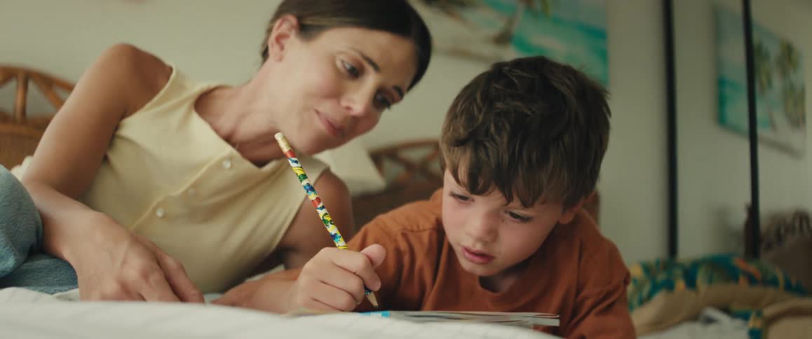 A loving mother smiles and talks to her son as he concentrates on drawing in a book while lying on the bed