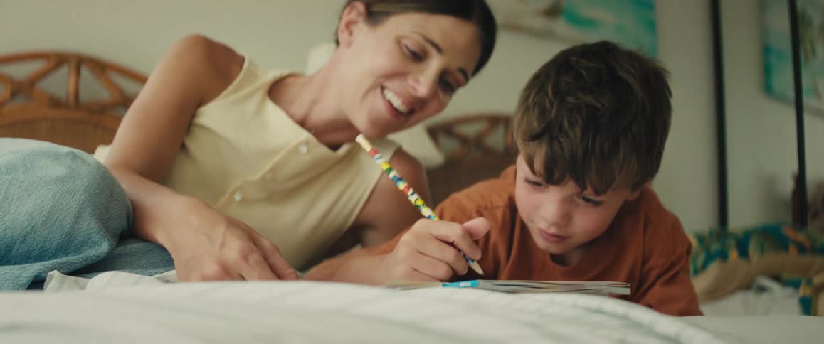 A loving mother smiles and talks to her son as he concentrates on drawing in a book while lying on the bed