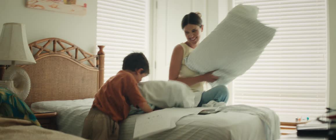 A smiling mother and her son laugh and play as they have a slow-motion pillow fight on the bed in a sunlit bedroom