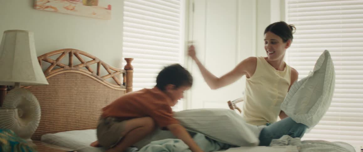 A smiling mother and her son laugh and play as they have a slow-motion pillow fight on the bed in a sunlit bedroom