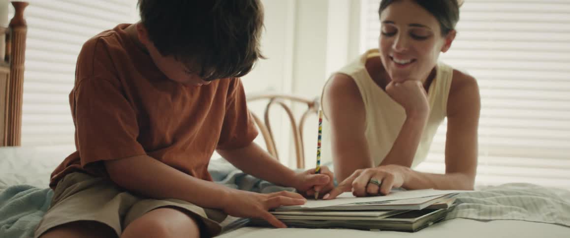 A young boy sits on a bed and concentrates on his drawing as his smiling mother watches and encourages him