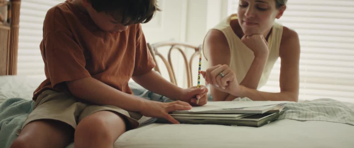 A young boy sits on a bed and concentrates on his drawing as his smiling mother watches and encourages him