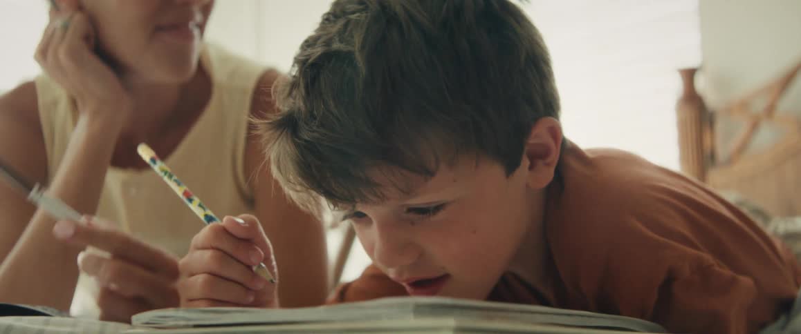A young boy intently practices writing in his notebook while his mother lovingly assists him with his homework
