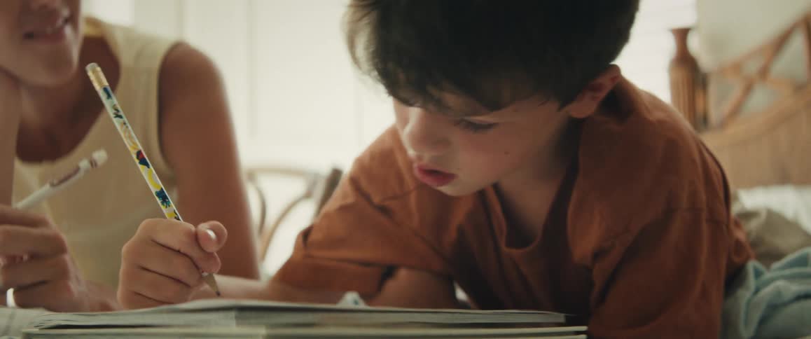 A young boy intently practices writing in his notebook while his mother lovingly assists him with his homework