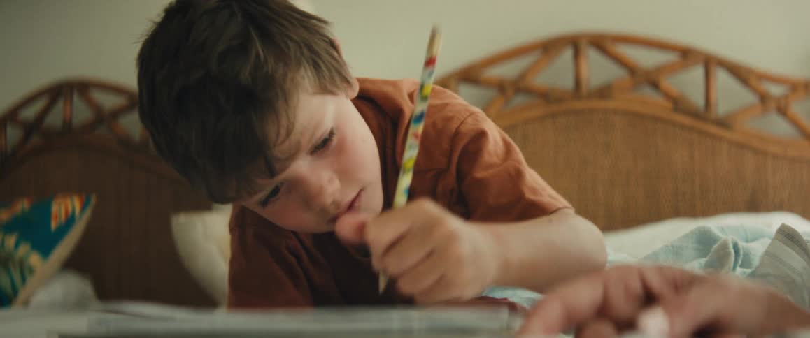 An intimate close-up shot captures a young boy concentrating intently as he writes in a notebook while lying on his bed