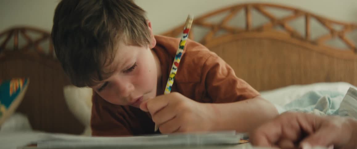 An intimate close-up shot captures a young boy concentrating intently as he writes in a notebook while lying on his bed