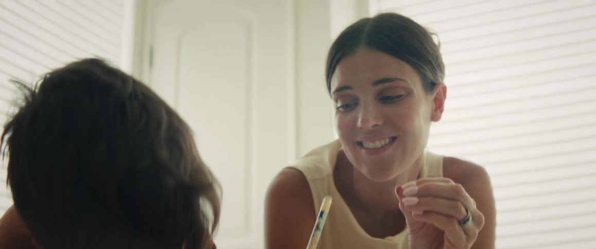 Close up of a proud mother smiling and lovingly watching her son as he draws at a table indoors