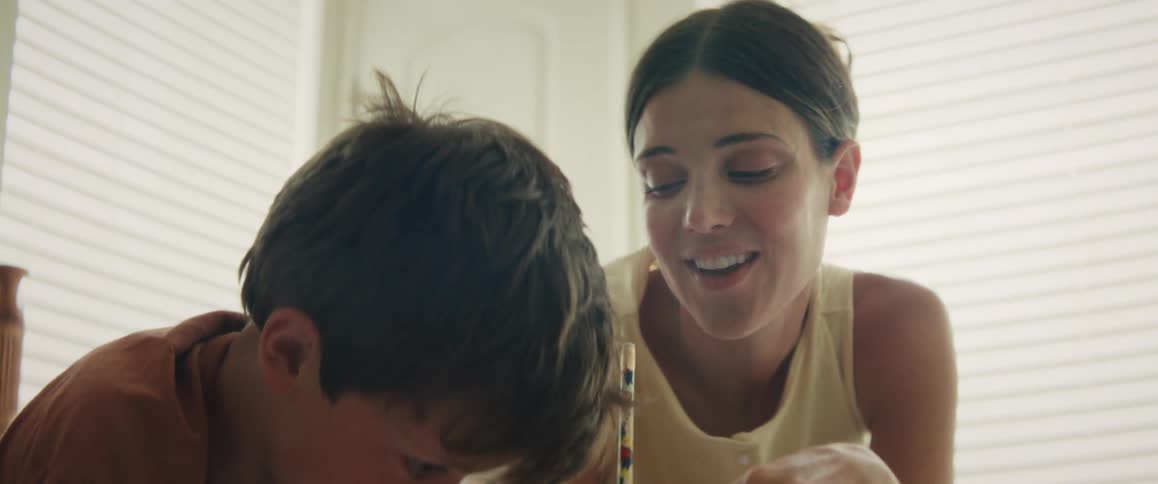 Close up of a proud mother smiling and lovingly watching her son as he draws at a table indoors